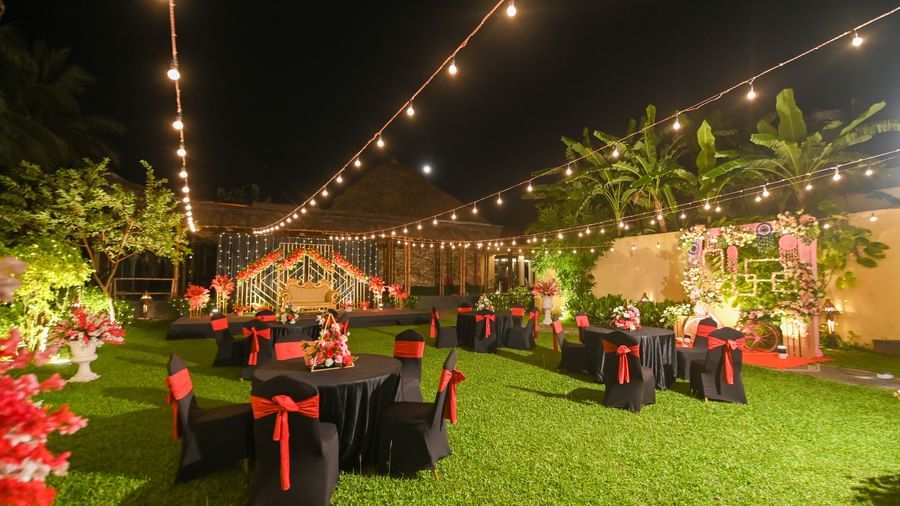 A night time view of a neatly trimmed lawn space at Vedic Village Spa Resort, featuring warm decorative lightsand black banquet chairs with red bows.
