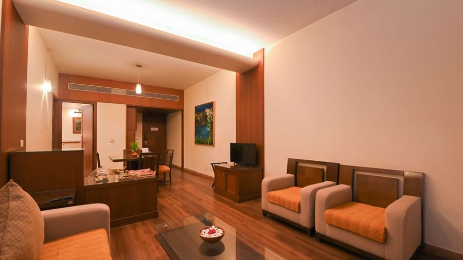 Corner view of the living room in the One-Bedroom Suite at Vedic Village Spa Resort featuring wooden flooring, a glass-topped table, plush couches, wooden furniture, dining area, and flat-screen TV.