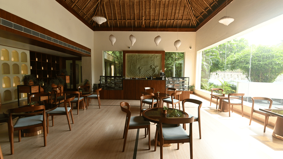 An open air restaurant seating area at Vedic Village Spa Resort Kolkata featuring wooden chairs and tables thatched roof and lush greenery visible through large open windows.