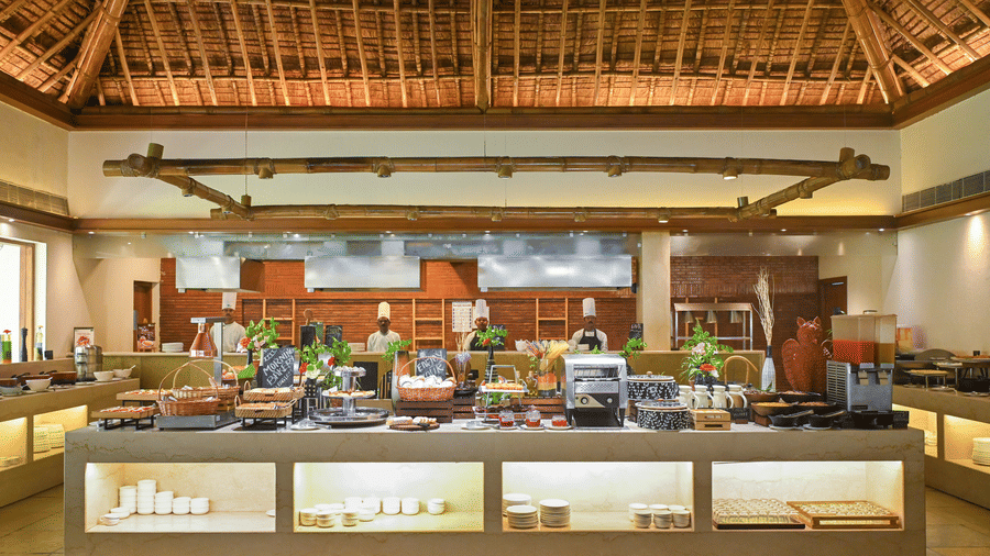Restaurant buffet spread at Vedic Village Spa Resort Kolkata with a long counter displaying multiple food dishes under a high exposed wooden thatched roof ceiling.