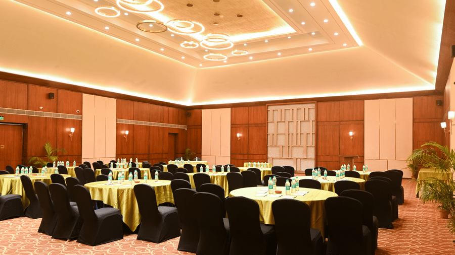 Banquet hall at Vedic Village Spa Resort Kolkata with round tables, yellow table linen, black chairs, wooden wall panelling, and warm decorative ceiling lighting.
