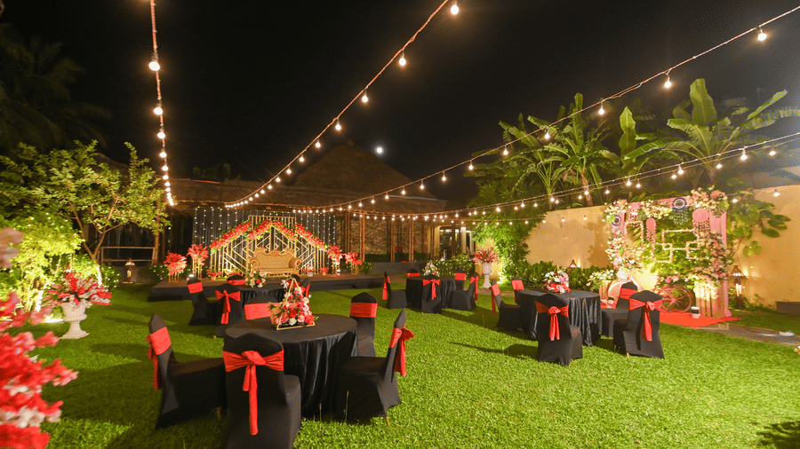 Outdoor garden event setup at Vedic Village Spa Resort Kolkata with fairy string lights, black chairs with red sashes, round tables, and warm ambient night lighting.