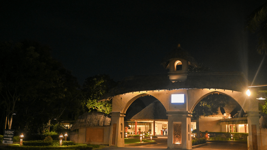 Illuminated resort gateway arch at Vedic Village Spa Resort Kolkata with warm lighting, decorative entrance structure, and a digital display board at night.