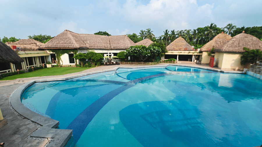 Outdoor swimming pool at Vedic Village Spa Resort Kolkata surrounded by thatched roof cottages, lush palm trees, and manicured gardens under a bright blue sky.