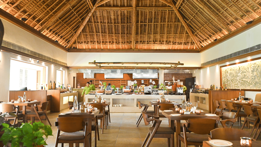 Resort restaurant at Vedic Village Spa Resort Kolkata with a high exposed wooden thatched roof, round dining tables, wooden chairs, and indoor plants with warm ambient lighting.