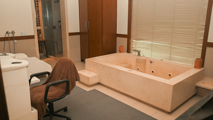 Spa treatment room at Vedic Village Spa Resort Kolkata featuring a wooden bathtub, massage chair, wooden cabinetry, and soft ambient lighting.