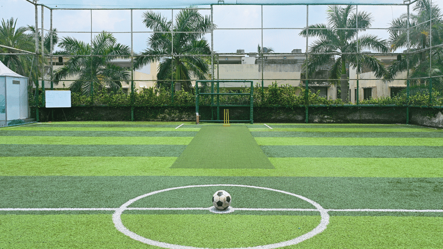 Synthetic football turf at Vedic Village Spa Resort Kolkata with a green playing surface, white centre circle markings, surrounding fencing, and palm trees in the background.
