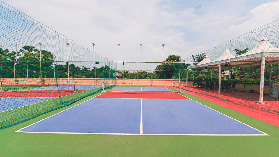 Outdoor tennis court at Vedic Village Spa Resort Kolkata with a blue and red playing surface, net, surrounding fencing, and floodlights under a clear blue sky.