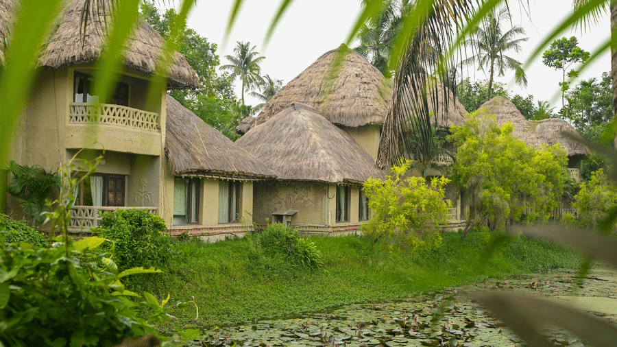 Thatched-roof Earth Villas at Vedic Village Spa Resort overlooking a water body, with lush green vegetation lining the perimeter of the natural surroundings.
