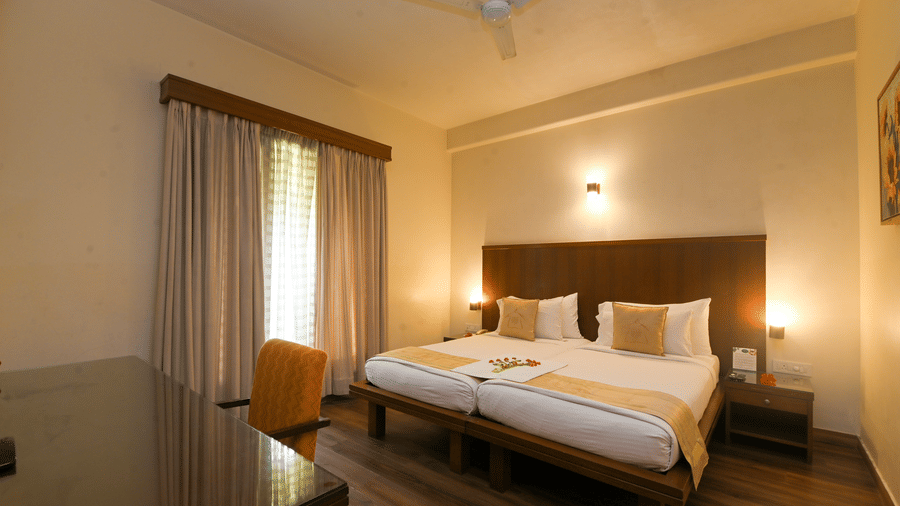 Warmly lit Superior Room at Vedic Village Spa Resort showcasing a double bed, wooden flooring, ceiling fan, and a work desk with chair for added convenience.