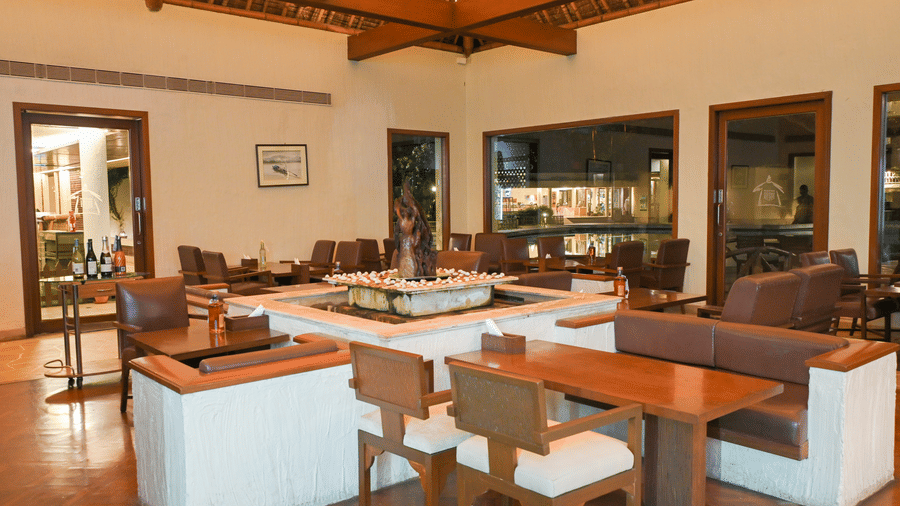 Villa living and dining area at Vedic Village Spa Resort Kolkata with a wooden dining table, sofa seating, framed artwork, and large windows with natural light.