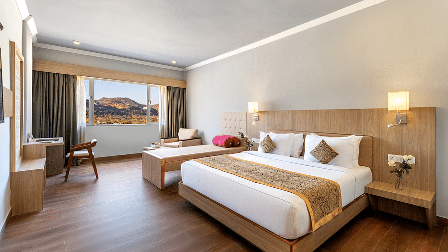 A bright hotel room with a neatly made bed, wooden furniture and a large window offering a view, with a minimalist design - Via Lakhela Resort & Spa, Kumbhalgarh