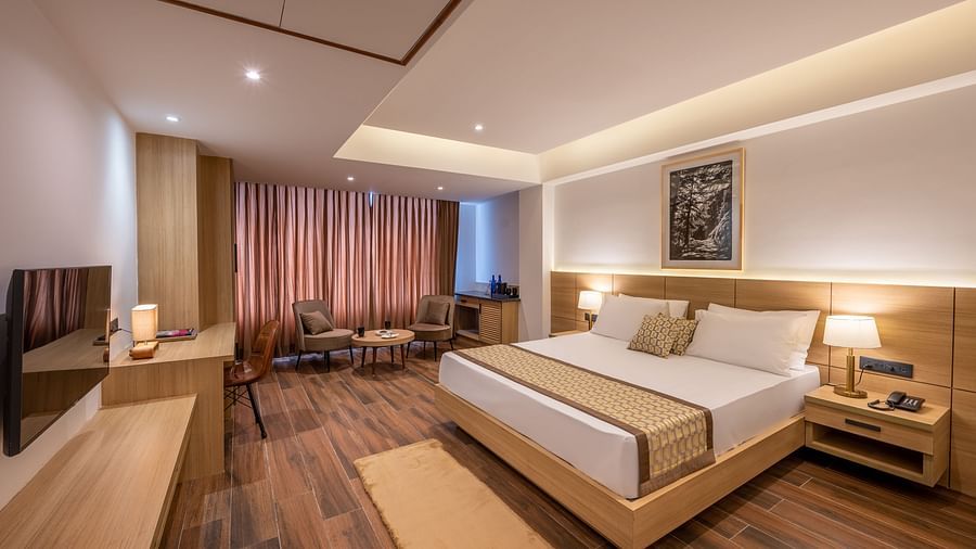 Full shot of Suite at Vishranti Hotel, Dehradun featuring a king bed with side tables, a TV, large window with curtains and bed lamps. 
