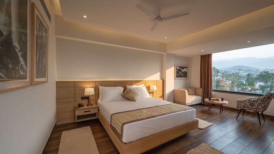 Full shot of Premium Room at Vishranti Hotel, Dehradun featuring a king bed with side tables, a TV, large window and bed lamps. 