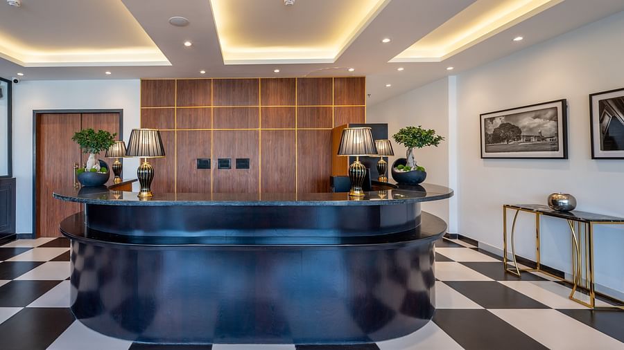  Reception area of Vishranti Hotel, Dehradun featuring a black reception desk with a curved design, a checkered black and white tiled floor, and a backdrop of wood-paneled walls.