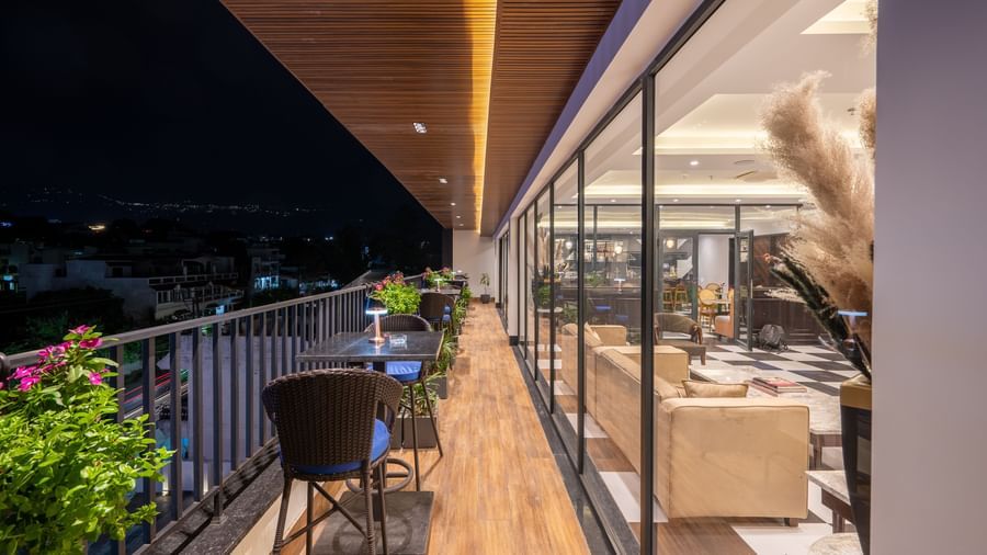 Overview of an outdoor dining area on a balcony, of The Spirit Tribe Restaurant at Vishranti Resort, Dehradun. The balcony features a dark-colored metal railing, a wooden floor, and a dining table with chairs.