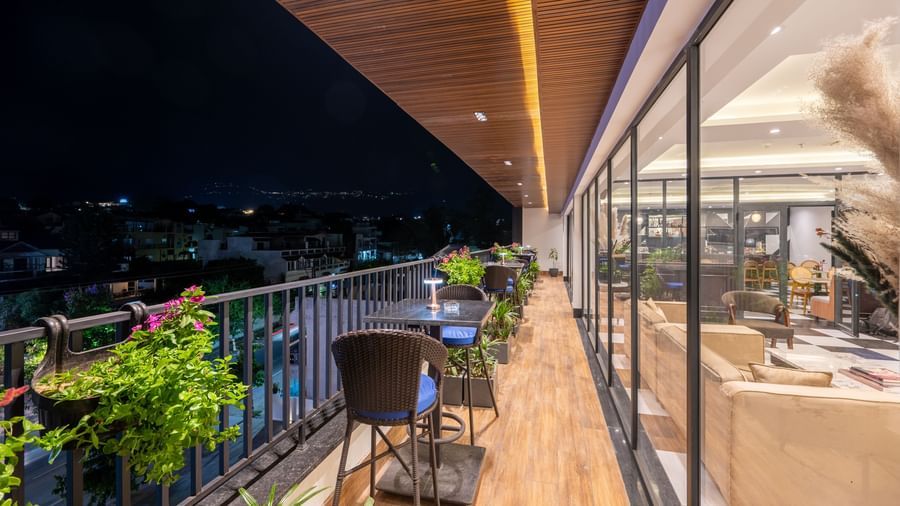 Overview of an outdoor dining area on a balcony, of The Spirit Tribe Restaurant at Vishranti Resort, Dehradun. The balcony features a dark-colored metal railing, a wooden floor, and a dining table with chairs.