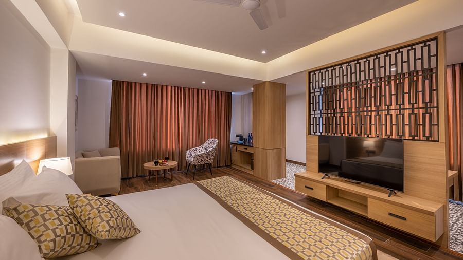 Close up shot of Suite at Vishranti Hotel, Dehradun featuring a king bed with side tables, a TV, large window with curtains and bed lamps. 