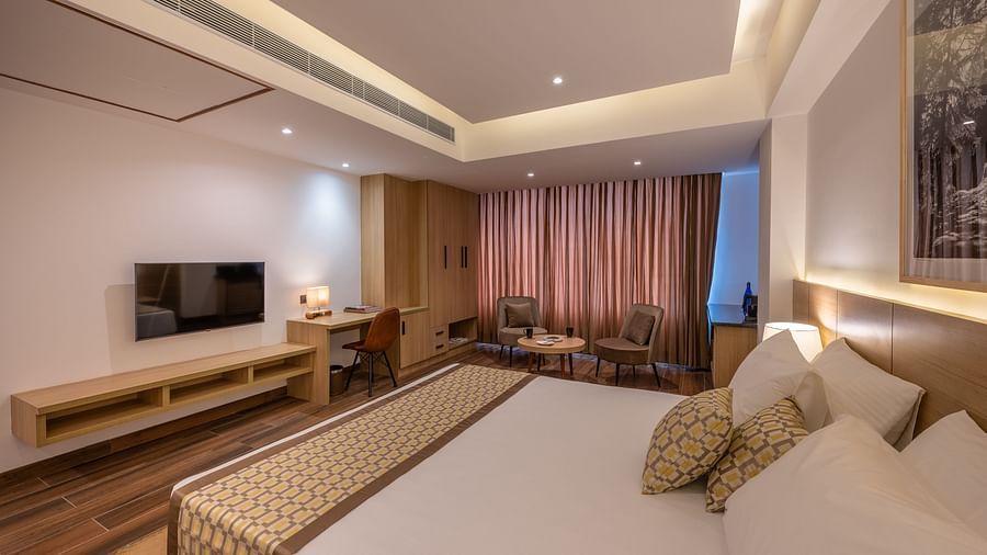 Side Shot of Premium Room at Vishranti Hotel, Dehradun featuring a king bed with side tables, a TV, large window and bed lamps. 
