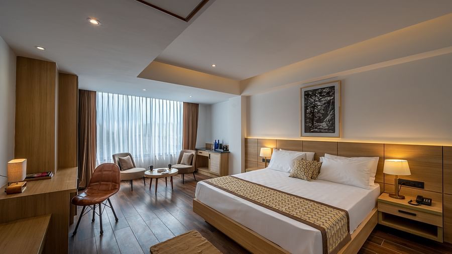 Shot of Premium Room at Vishranti Hotel, Dehradun featuring a king bed with side tables, a TV, large window and bed lamps. 