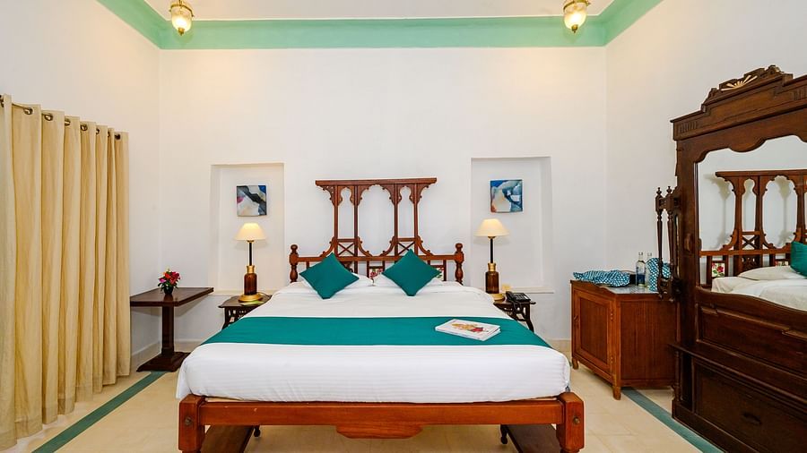 A bedroom with a wooden bed decorated with pillows and a bed runner, side lamps, framed art, and a wooden cabinet on the side. - Tijara Fort-Palace - 19th Century, Alwar