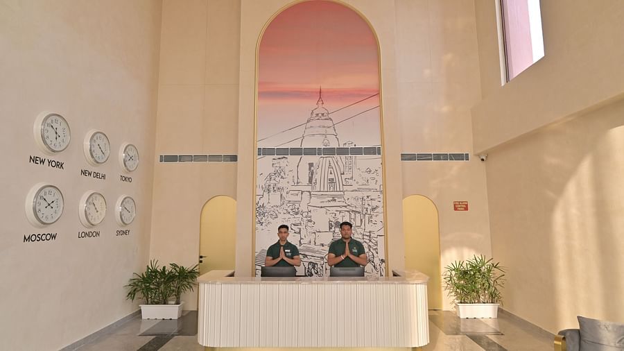 A reception area with two receptionists behind a white desk, clocks displaying different time zones on the wall, and plants on either side. - Clarks Safari Haridwar