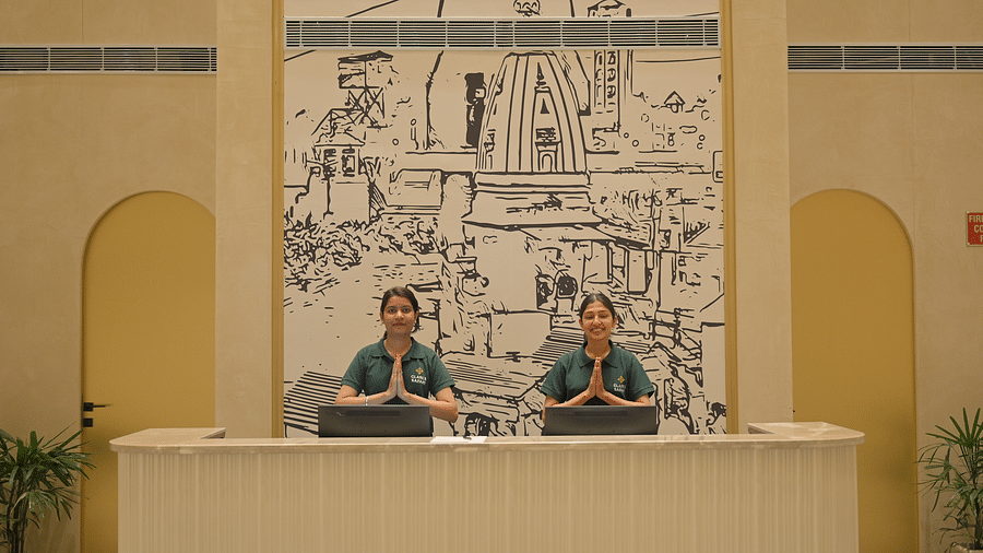 Two people standing behind a reception desk with a large monochrome drawing on the wall behind them. - Clarks Safari Haridwar