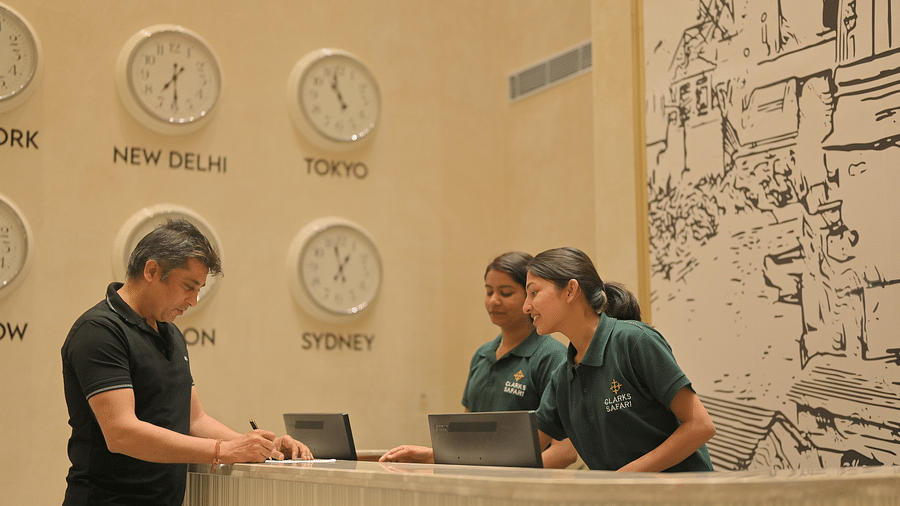 A man checking in at the front desk with several clocks on the wall showing different time zones. - Clarks Safari Haridwar
