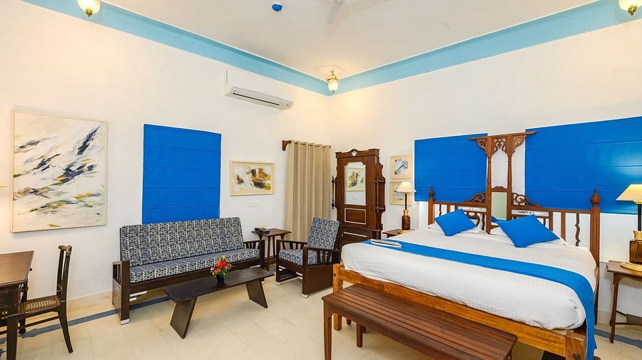 A spacious room featuring a wooden four-poster bed, blue accents, a gray couch, a dark wood table, and a decorative painting, offering a blend of comfort and style - Tijara Fort-Palace - 19th Century, Alwar.