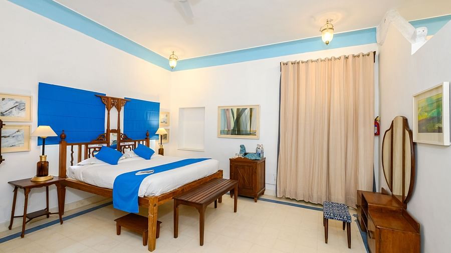 A bedroom with a wooden four-poster bed, a blue accent wall, two side tables, a wooden dresser with a mirror, and a beige curtain - Tijara Fort-Palace - 19th Century, Alwar.