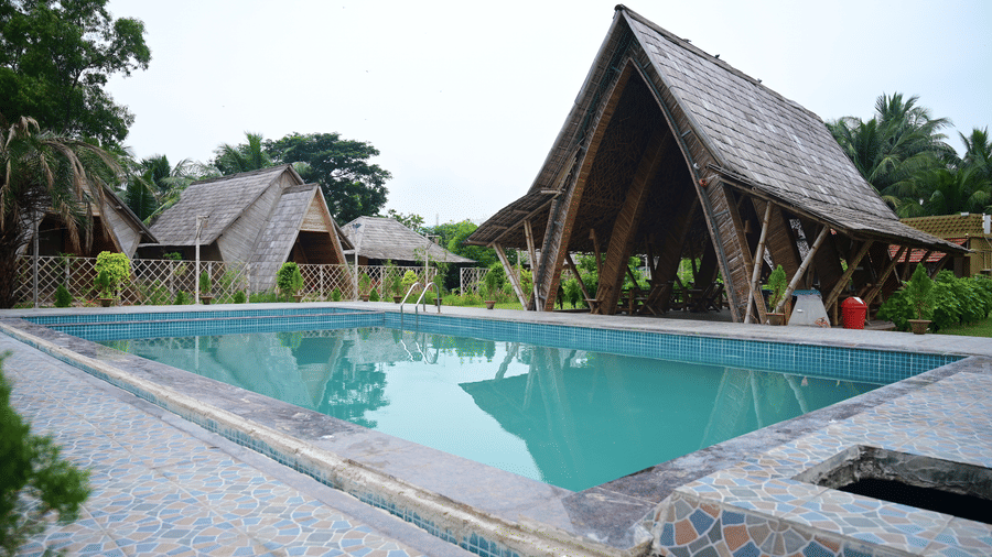 An outdoor shot of the swimming pool at Pipul Odi Art Museum Resort, featuring clear blue water and surrounded by paved decking, with A-frame cottages visible in the background.