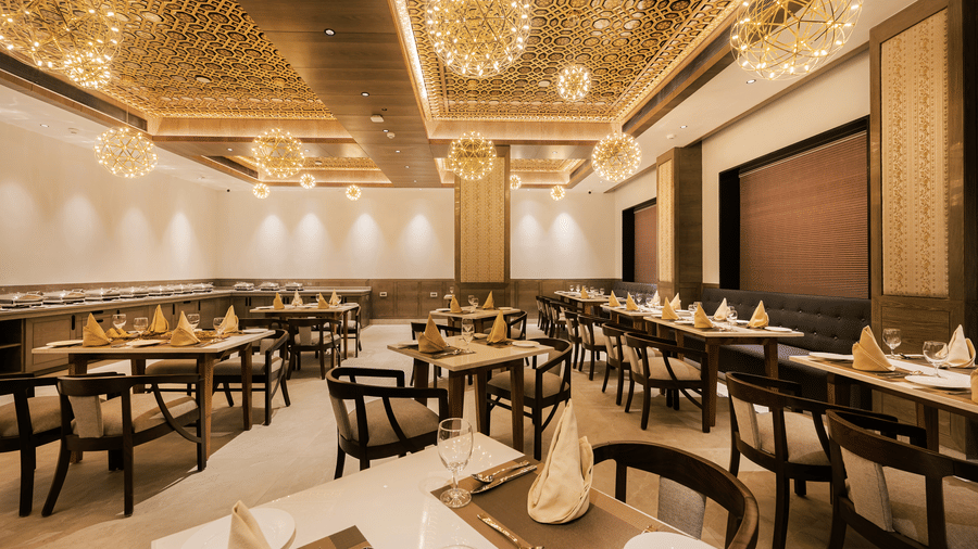 A spacious restaurant dining hall featuring wooden tables, black chairs, and modern gold-tone chandeliers at Ananta Express, Katra.