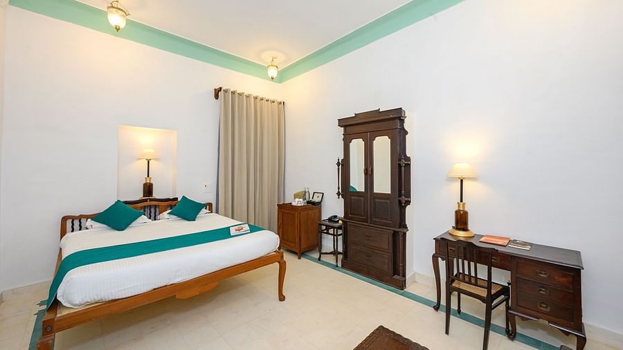 A room with a wooden bed, teal accents, a dark wood armoire, a desk with a chair, and two lamps, showcasing the room's simple elegance - Tijara Fort-Palace - 19th Century, Alwar.
