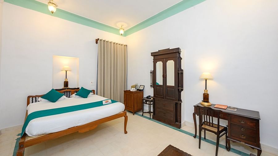 A room with a wooden bed, teal accents, a dark wood armoire, a desk with a chair, and two lamps, showcasing the room's simple elegance - Tijara Fort-Palace - 19th Century, Alwar.
