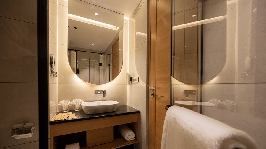 A contemporary bathroom vanity with a white vessel sink, a large mirror, and glass partitions at Ananta Express, Katra.