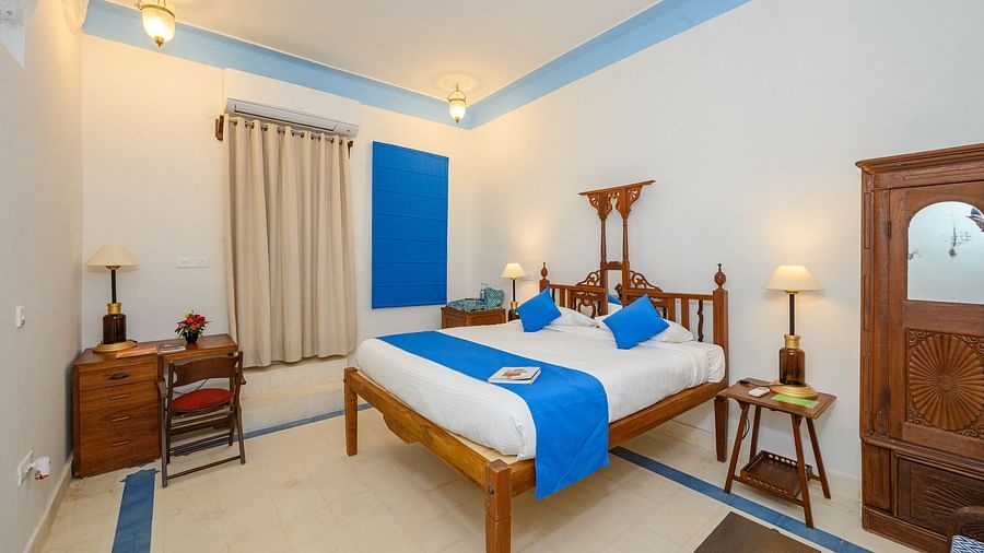 A bedroom with a wooden bed decorated with pillows and a runner, a wooden cabinet and a bed side table with a lamp on it. - Tijara Fort-Pal-Palace - 19th Century, Alwar