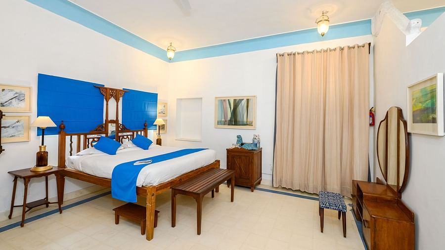 A bedroom with a wooden four-poster bed, a blue accent wall, two side tables, a wooden dresser with a mirror, and a beige curtain - Tijara Fort-Palace - 19th Century, Alwar.
