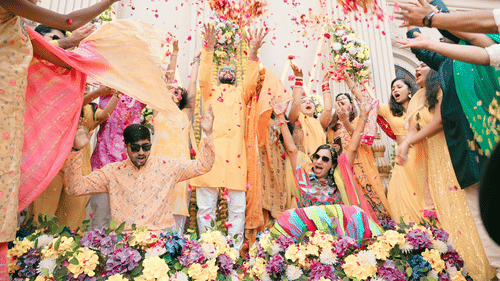 A couple in bridal shower decorated with flowers and garlands. There are people around pouring buckets filled with milk and flower petals.