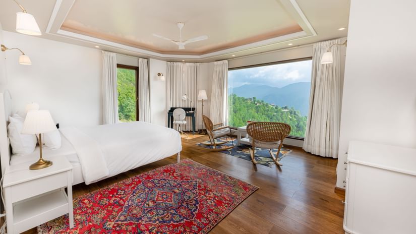 Bright bedroom at The Manor Luxury Apartments, Shimla, featuring a plushy bed, elegant furnishings, large picture windows, and panoramic views of the surrounding green hills.