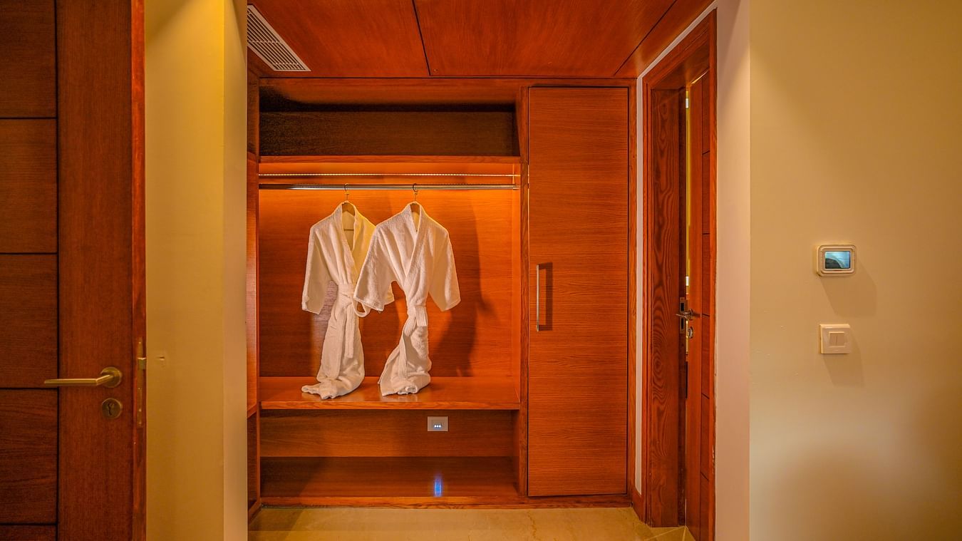 An open closet in a hotel room reveals hanging white robes and wooden shelves with folded towels. The closet is well-lit, and the interior is finished with wood - Ananta Spa and Resort, Pushkar