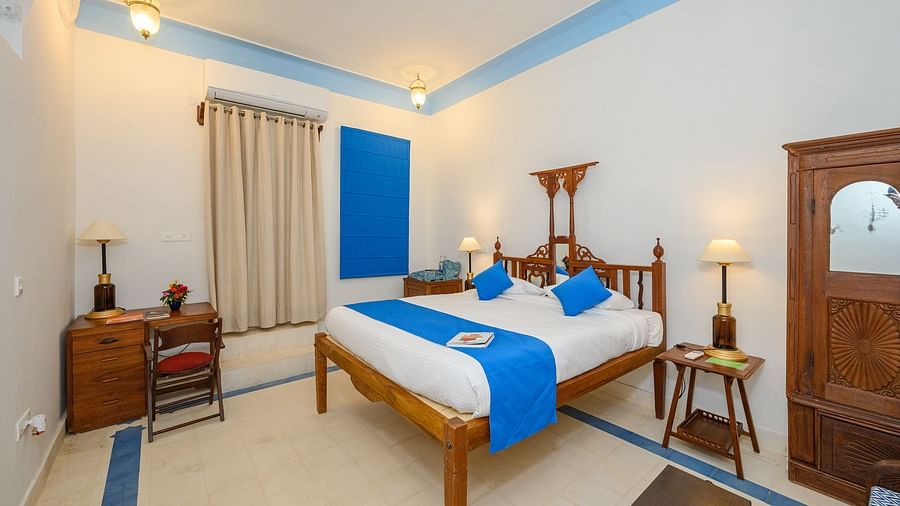 A bedroom with a wooden bed decorated with pillows and a runner, a wooden cabinet and a bed side table with a lamp on it. - Tijara Fort-Pal-Palace - 19th Century, Alwar