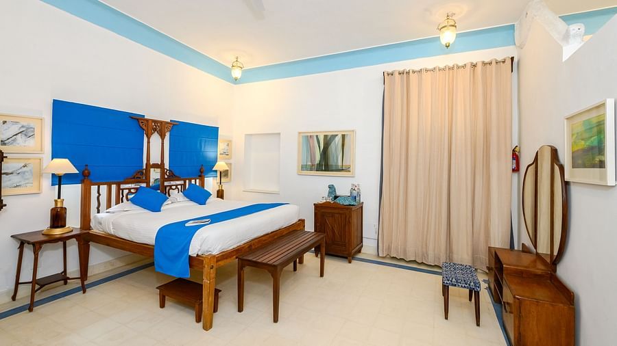 A bedroom with a wooden four-poster bed, a blue accent wall, two side tables, a wooden dresser with a mirror, and a beige curtain - Tijara Fort-Palace - 19th Century, Alwar.