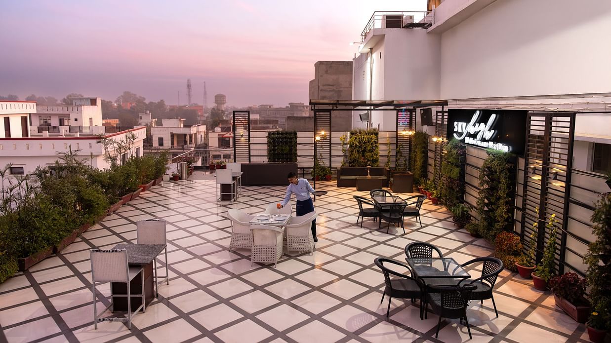  A rooftop terrace offering a stunning view of the city, with comfortable outdoor seating and a tranquil setting with a  man  fixing things on the table - Clarks Inn Express, Near Jammu Bari Brahmana Railway Station