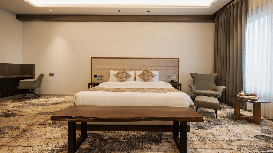 A guest room featuring a king-sized bed, a wooden bench, and a work desk area with an armchair at Ananta Express, Katra.
