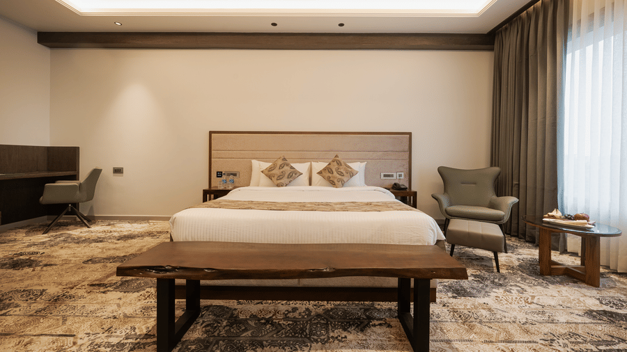 A guest room featuring a king-sized bed, a wooden bench, and a work desk area with an armchair at Ananta Express, Katra.