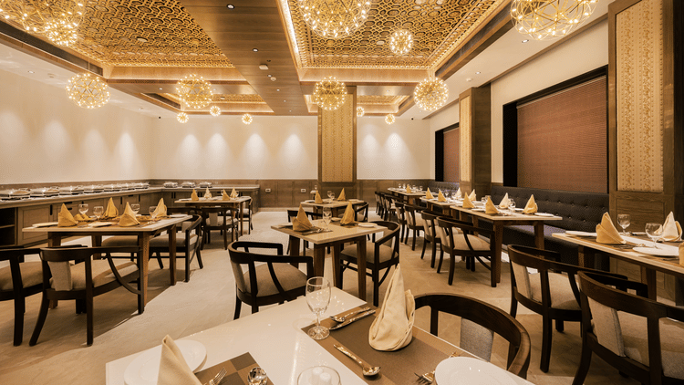 A spacious restaurant dining hall featuring wooden tables, black chairs, and modern gold-tone chandeliers at Ananta Express, Katra.