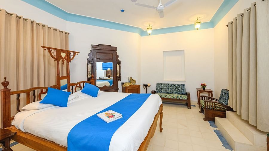 A bedroom with a wooden bed decorated with pillows and a bed runner, a seating area,and a cupboard in the background. - Tijara Fort-Palace - 19th Century, Alwar