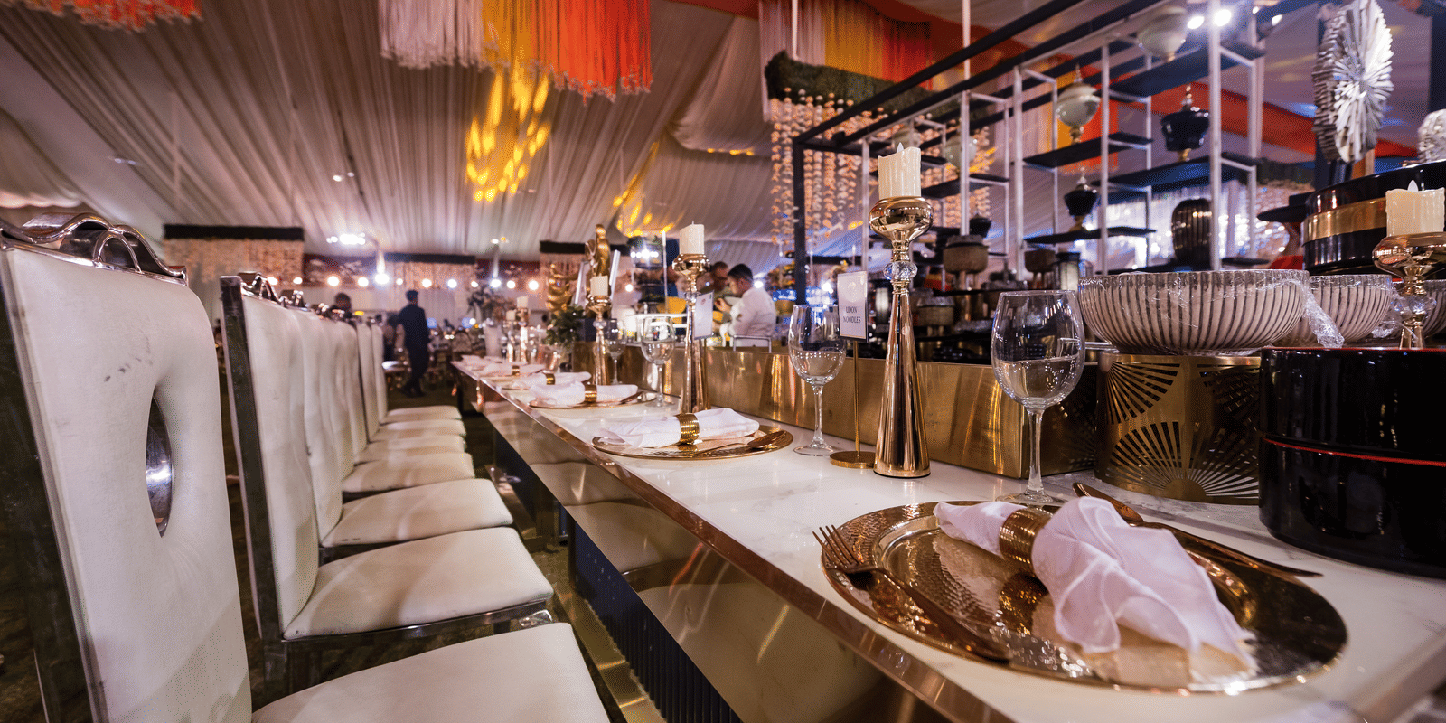 A long, formal buffet setup with catering dishes and serving equipment under event lighting in a temporary tent at Hotel Hukam's Lalit Mahal.