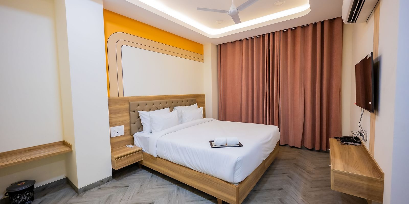 A cosy hotel room with soft lighting, a bed, curtains, and a TV mounted on the wall.