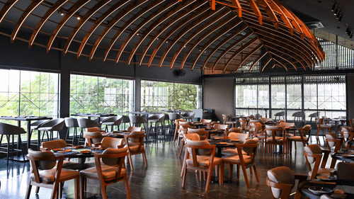 Contemporary restaurant dining room at Hotel Daspalla, Hyderabad featuring curved illuminated wooden ceiling, wooden chairs, dark tables, and floor-to-ceiling windows with garden views.